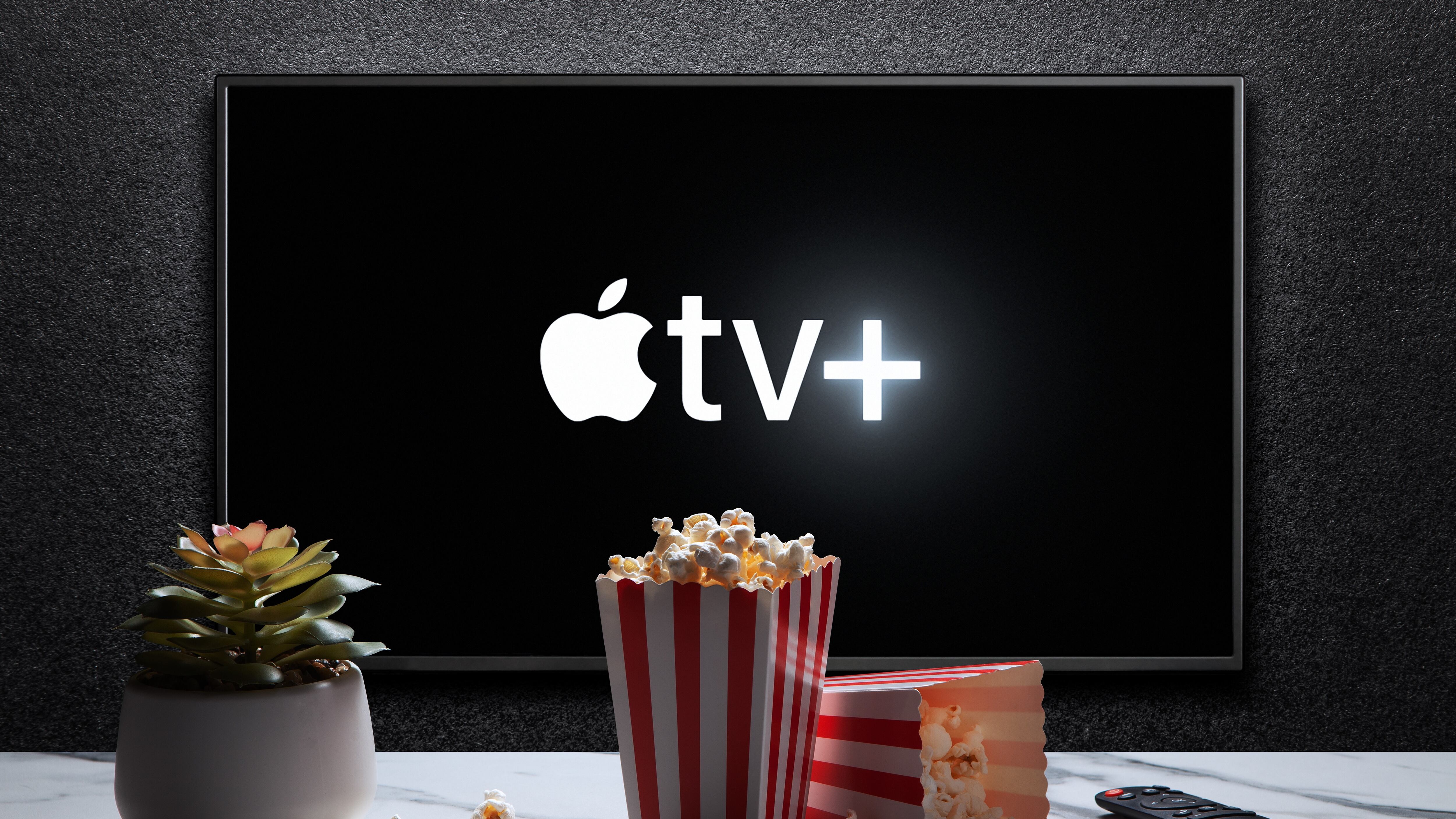 Apple TV plus logo on TV with popcorn boxes, remote control and plant on the table. Apple TV plus is an American subscription streaming service.